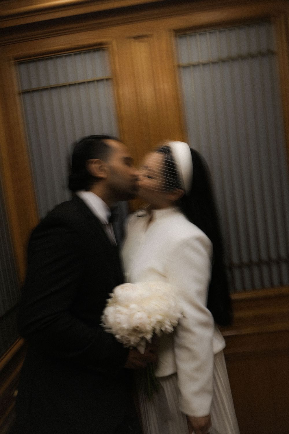 Bride and groom sharing a kiss at their wedding, bride holding white peony bouquet wearing veil and white jacket.