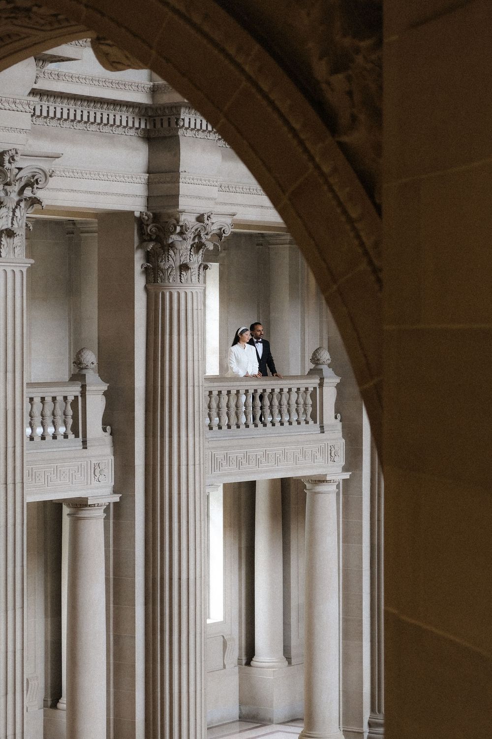 Bride and groom stand on ornate balcony surrounded by classical columns in grand architectural building.