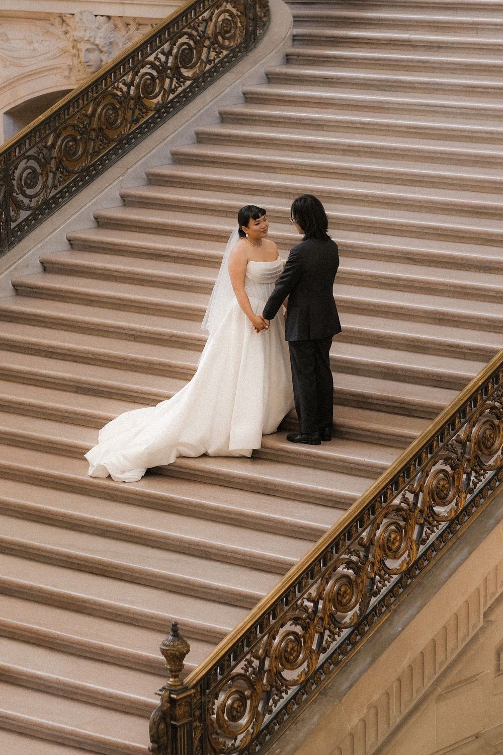 Bride and groom hold hands on grand marble staircase with ornate gold railings in elegant wedding venue.