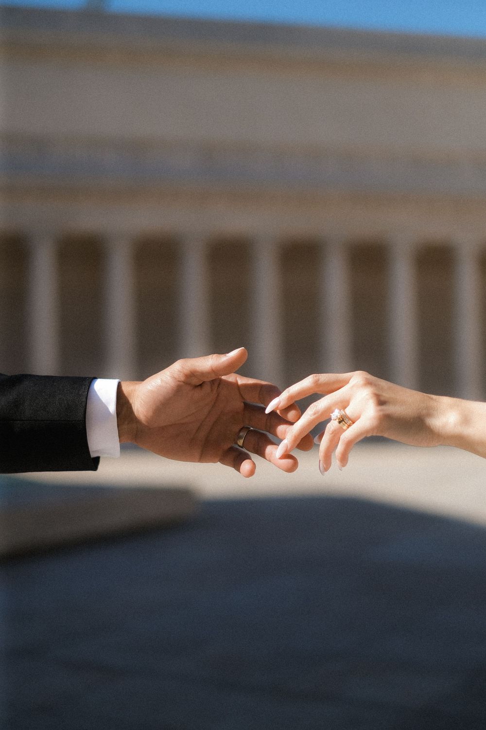 Interracial couple touching hands with wedding rings, classical columns building in background.