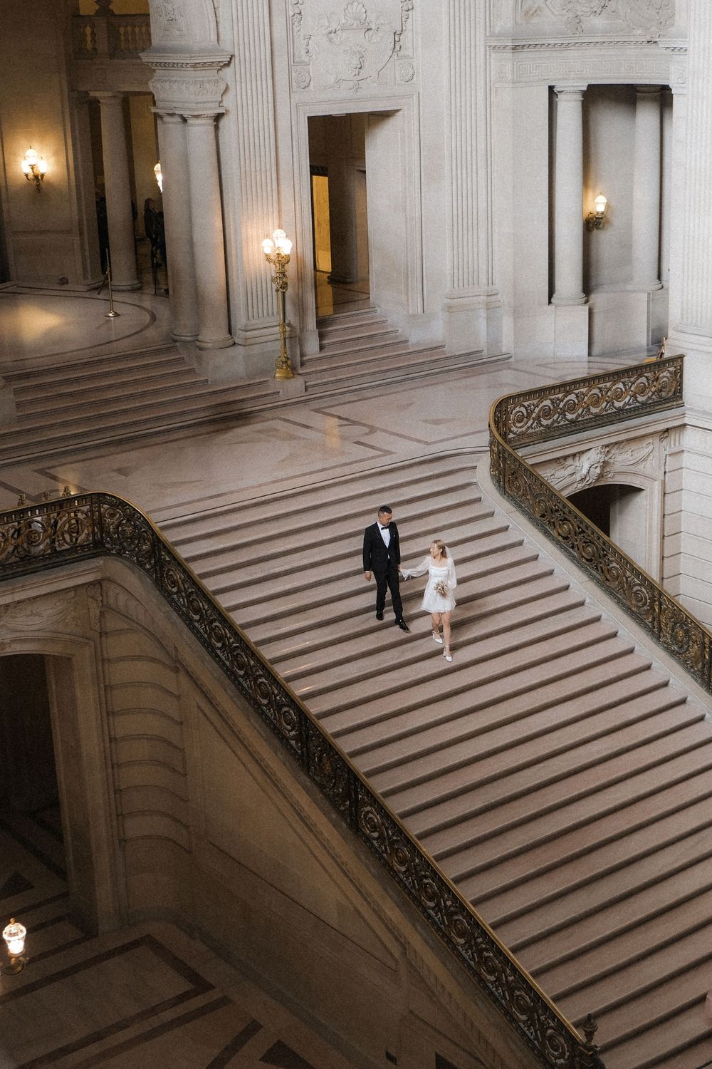Bride and groom walking down grand staircase at San Francisco City Hall during elegant wedding ceremony.