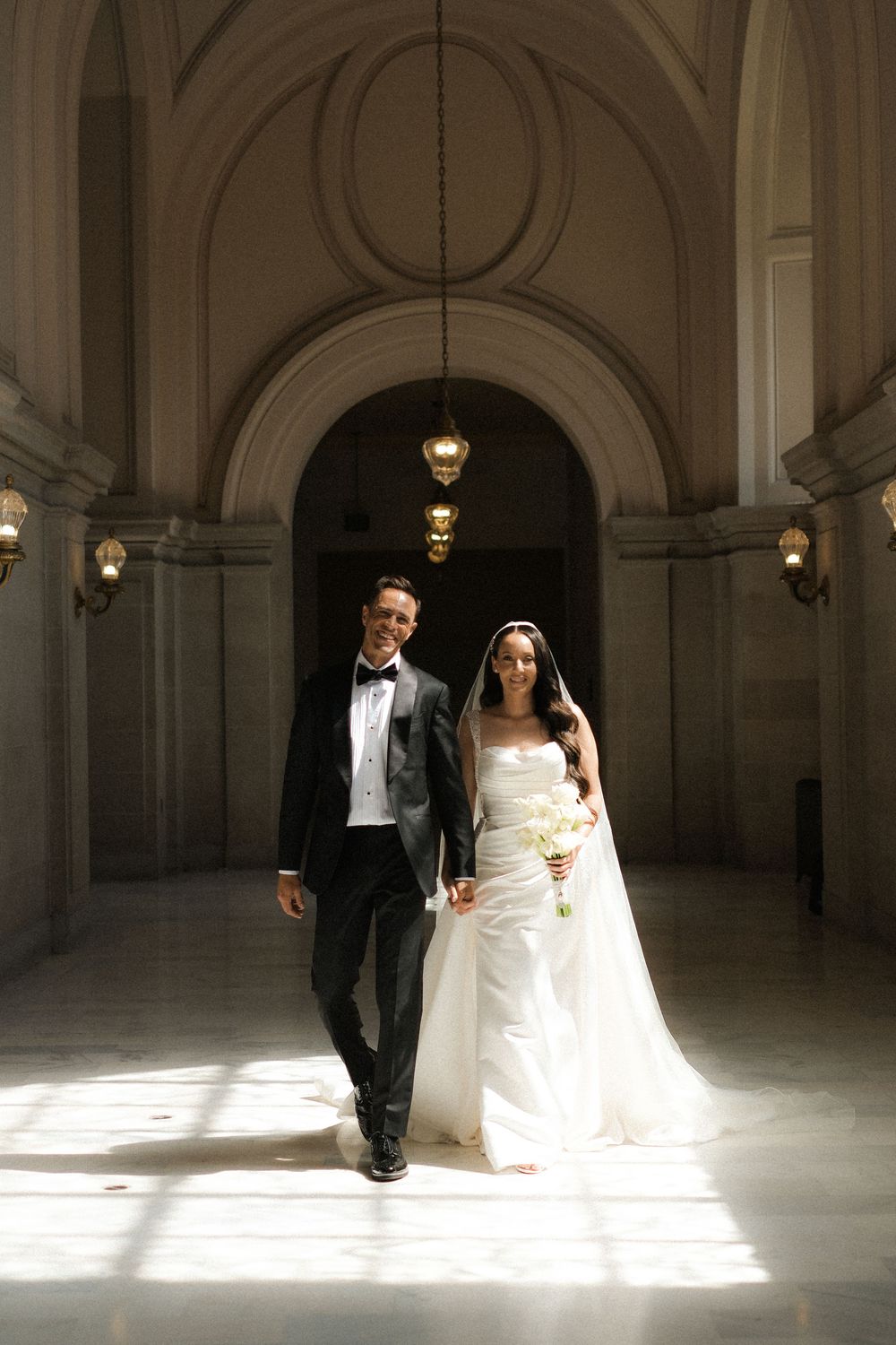 Bride and groom walk hand in hand through sunlit arched corridor of San Francisco City Hall.