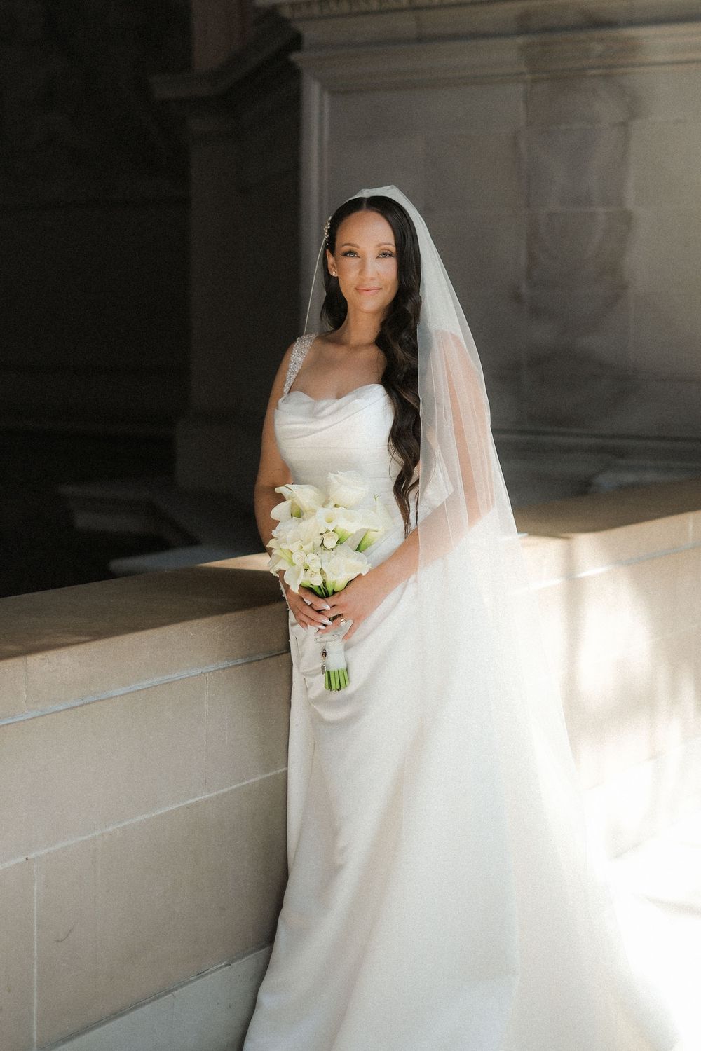 Elegant bride in white satin gown and veil holds white bouquet, posing gracefully against stone architecture in soft natural light.
