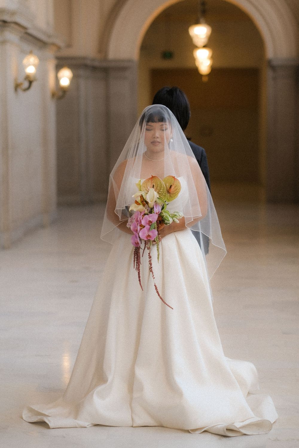 Bride in elegant white ballgown with veil holds colorful bouquet in grand hall with arched ceilings.