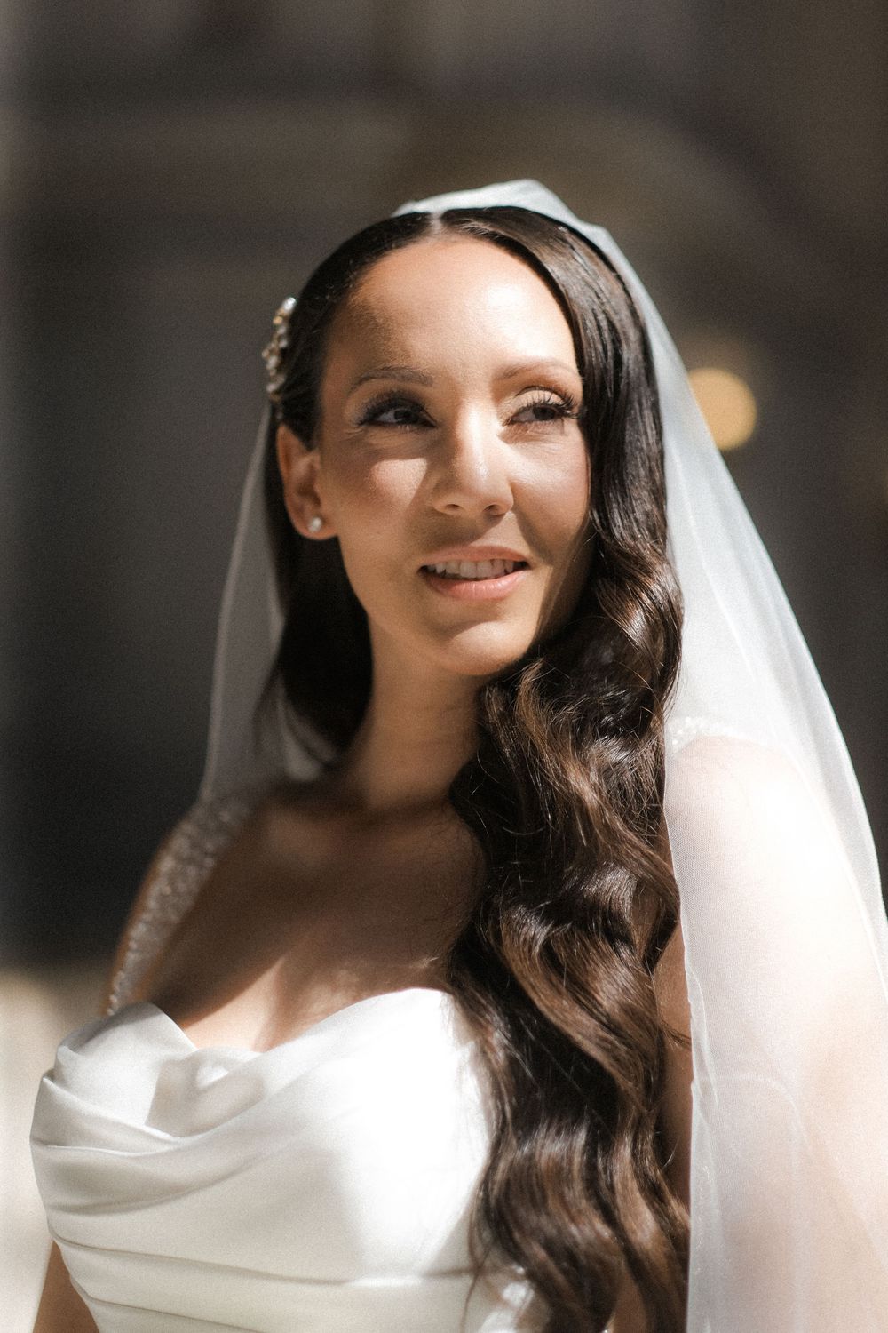 Radiant bride with long dark wavy hair and white veil smiles softly in strapless wedding gown.