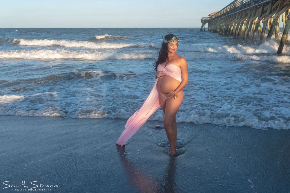 A maternity photo shoot on a beach at sunset with waves and a pier in the background.