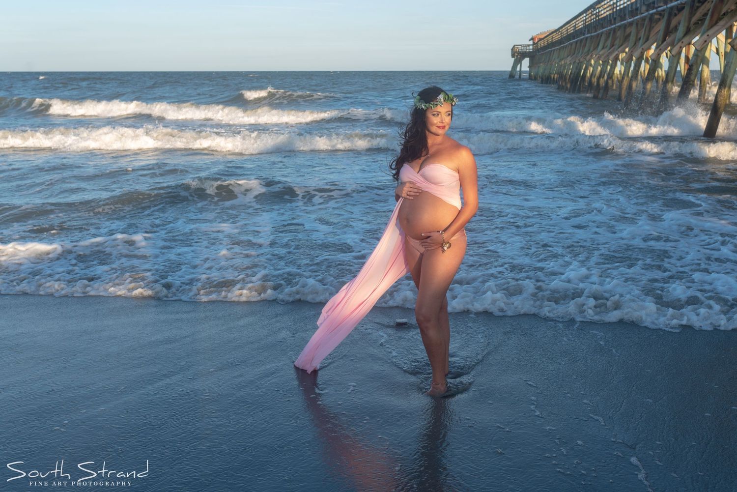 A maternity photo shoot on a beach at sunset with waves and a pier in the background.