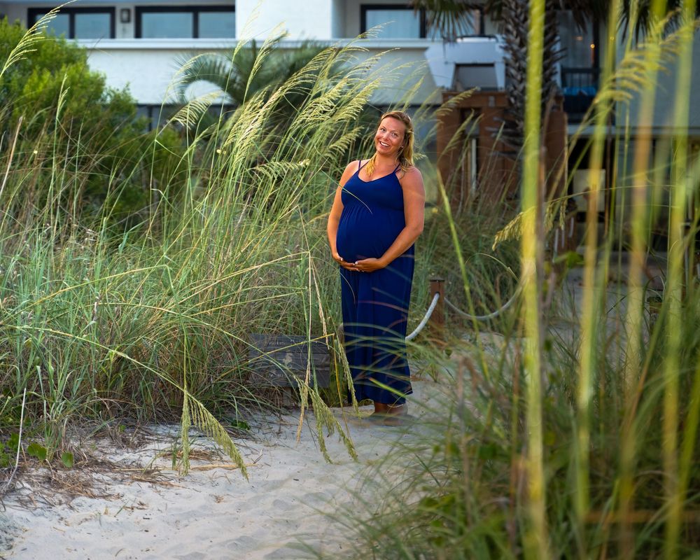 A person in a blue dress stands among tall beach grass with modern buildings visible in the background.