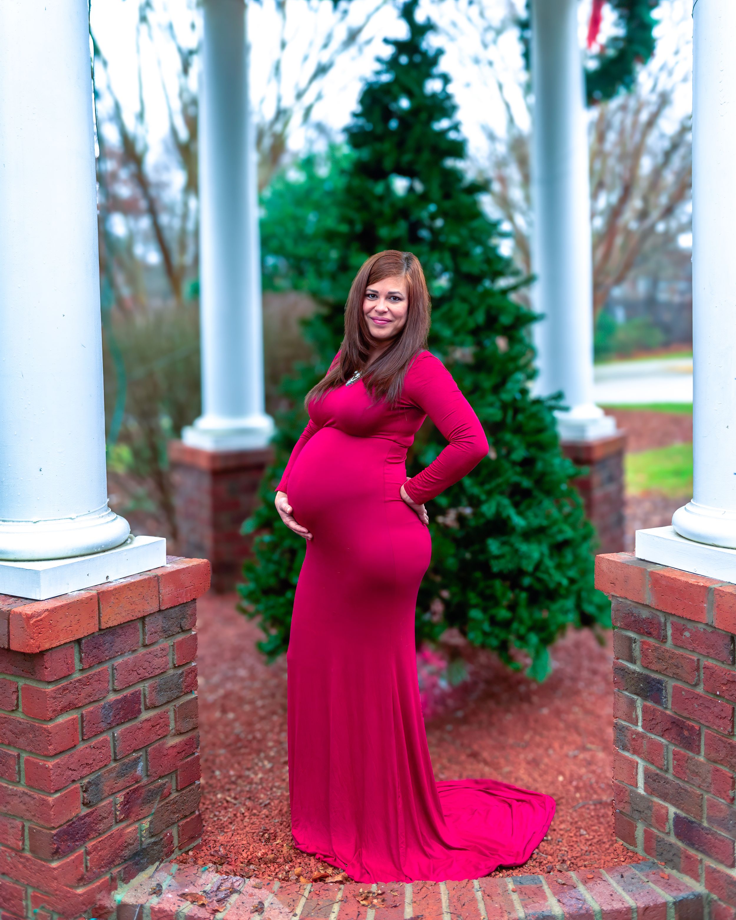 A woman in a flowing red dress poses gracefully between white columns on a brick porch with evergreen trees.