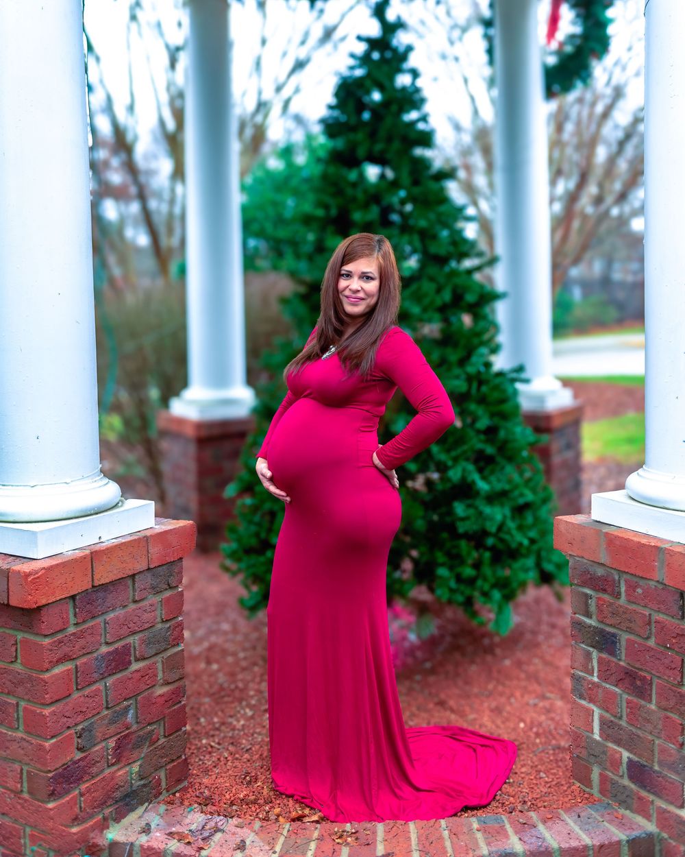A woman in a flowing red dress poses gracefully between white columns on a brick porch with evergreen trees.