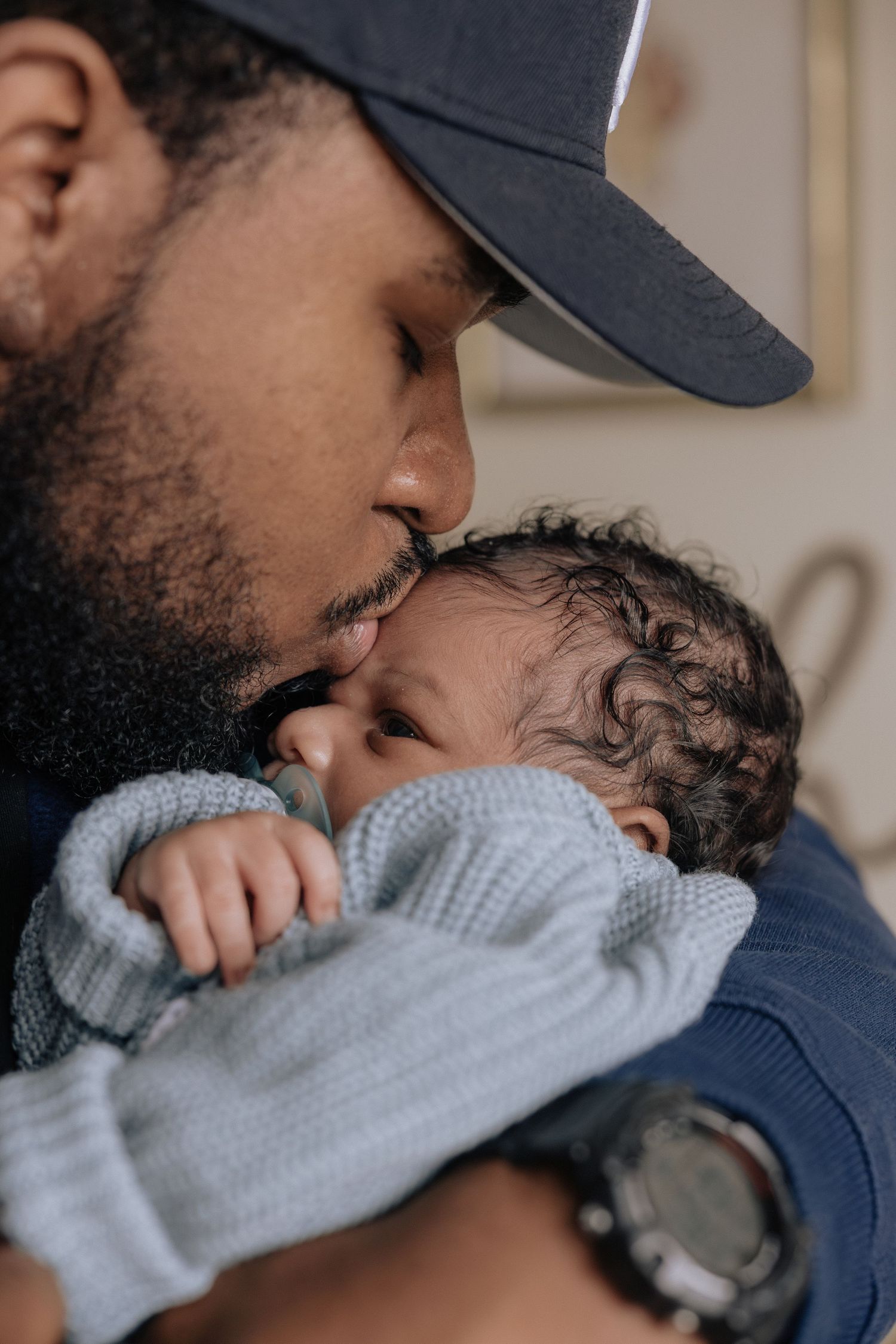 A parent wearing a blue baseball cap shares a tender moment with their baby wrapped in a soft gray blanket.