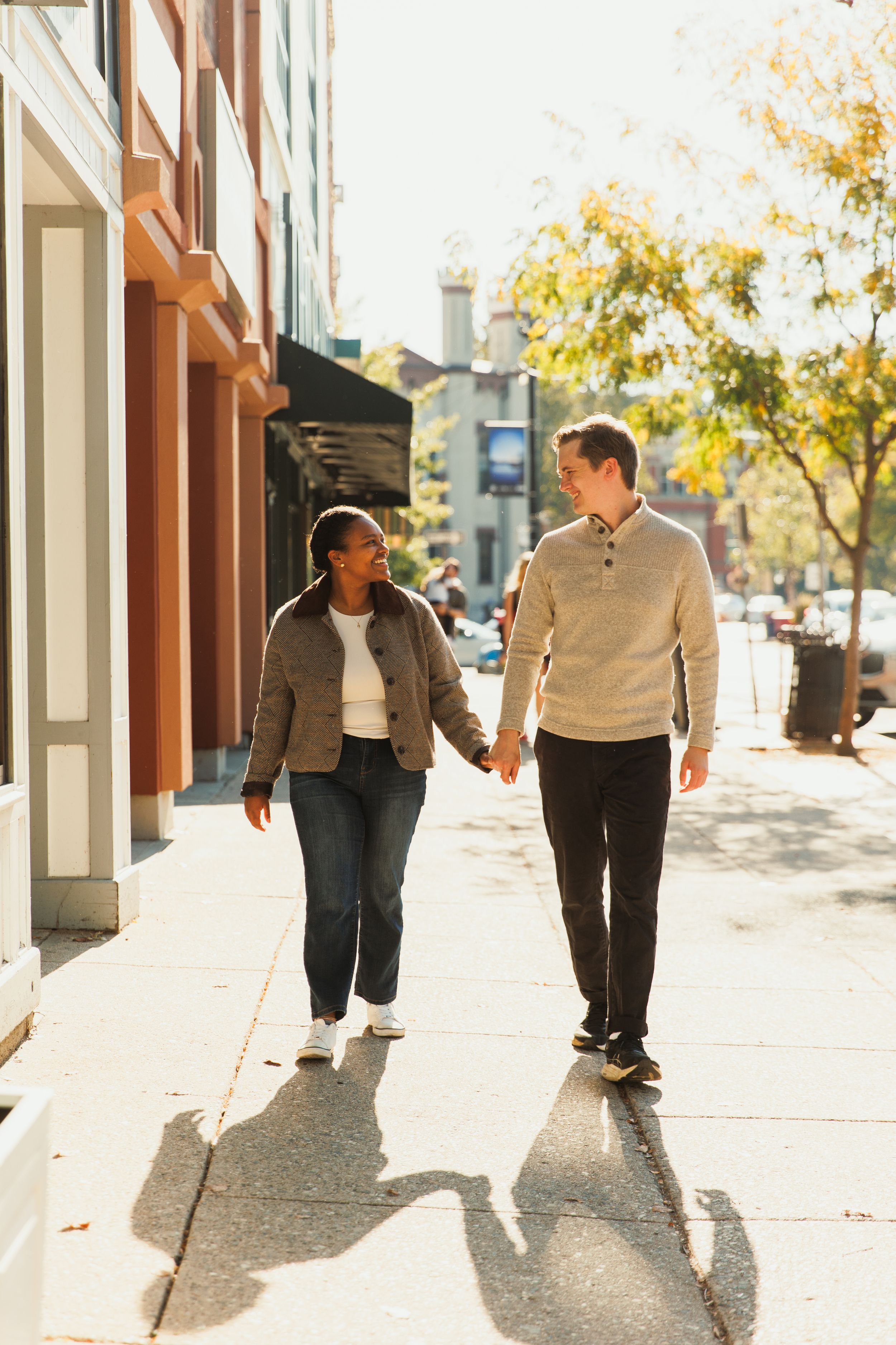 A couple holding hands while walking down a sunny sidewalk on an autumn day in the city.