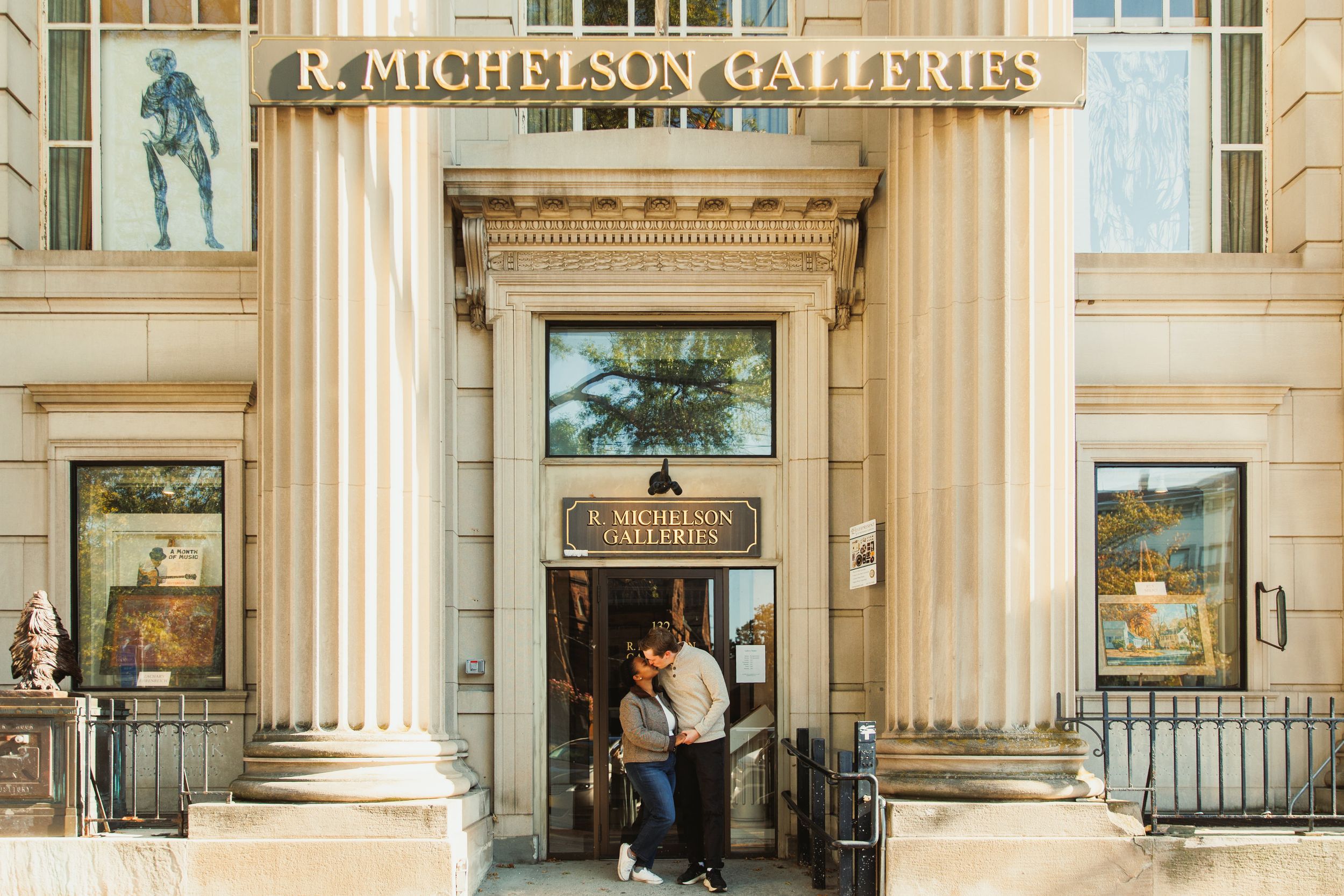 Art gallery entrance with classical columns and framed paintings displayed in elegant window cases along a stone facade.