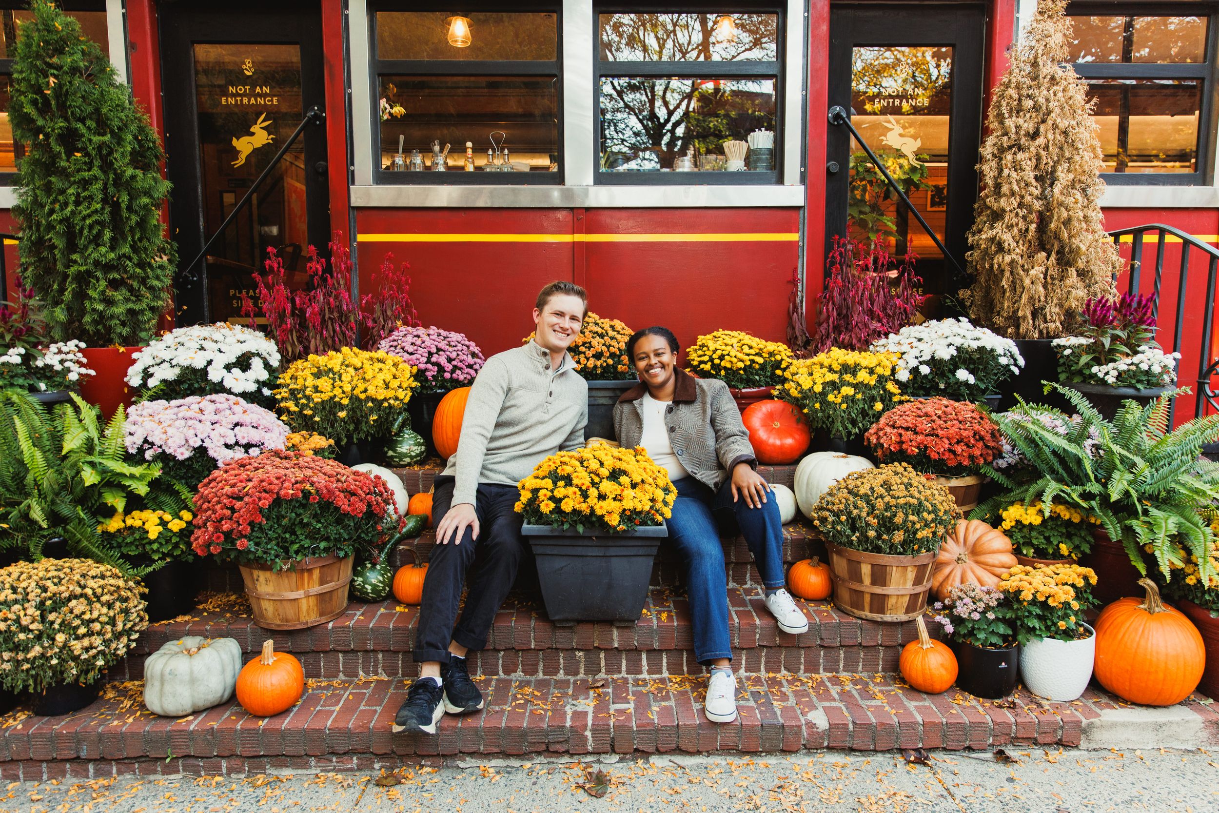 A cozy fall scene with people sitting among colorful mums, pumpkins and autumn decorations outside a red storefront.