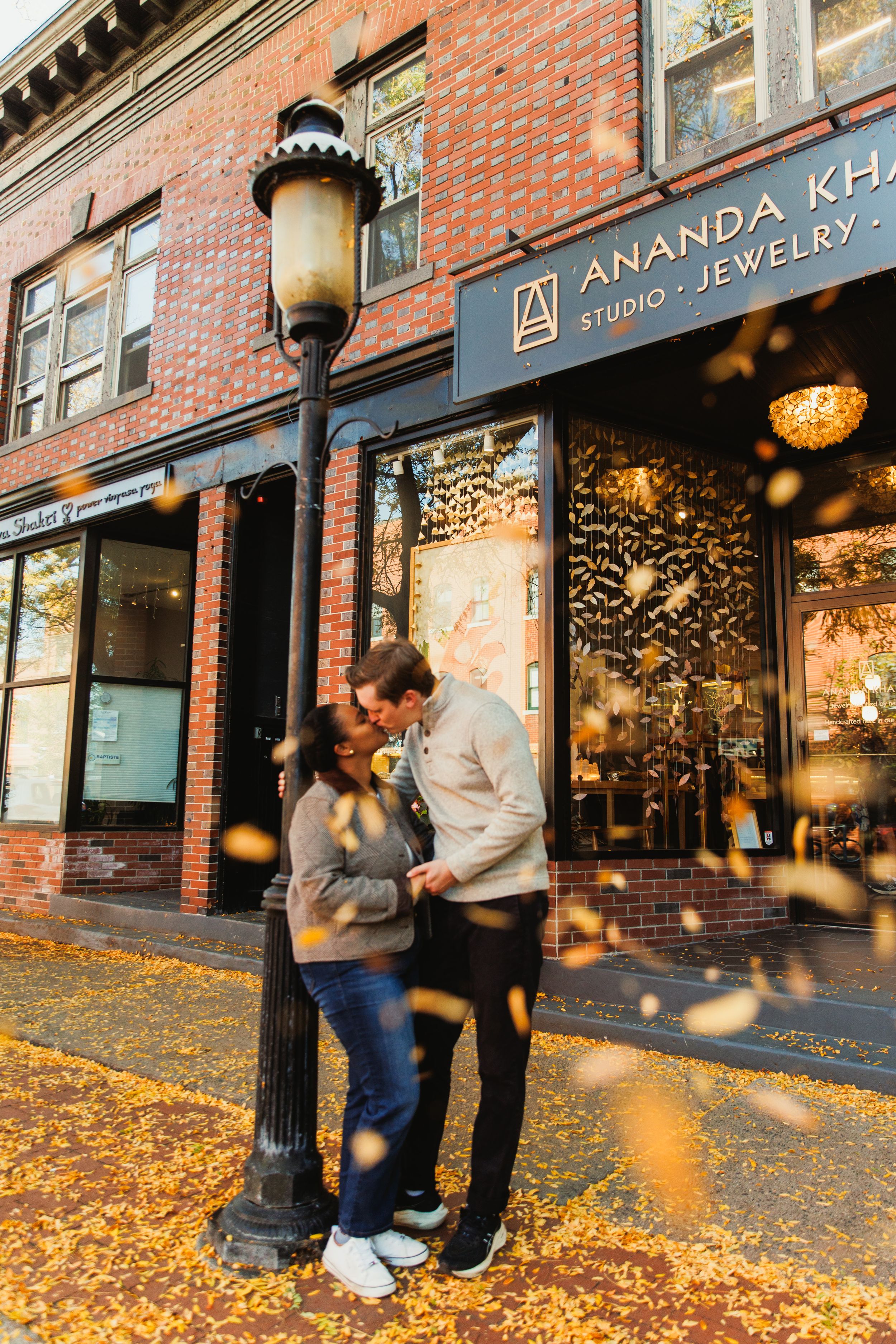 A couple embraces by a vintage lamppost outside a jewelry store on an autumn day with fallen leaves on the ground.