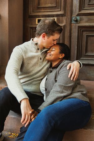 A couple sharing an intimate moment while sitting on wooden steps in casual sweaters and jeans.