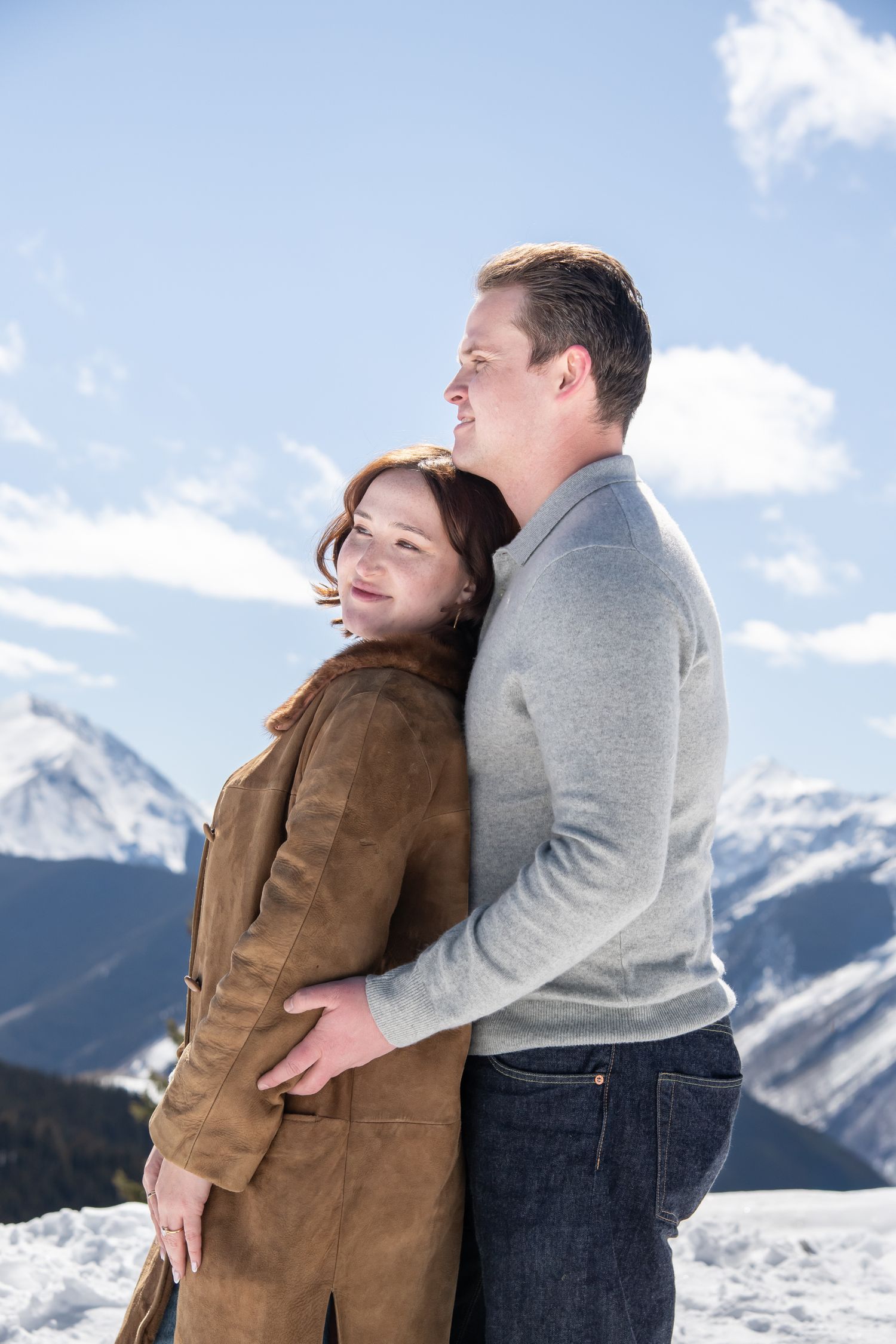 A couple embraces while enjoying a scenic mountain vista with snow-capped peaks and blue skies in the background in Aspen Colorado.