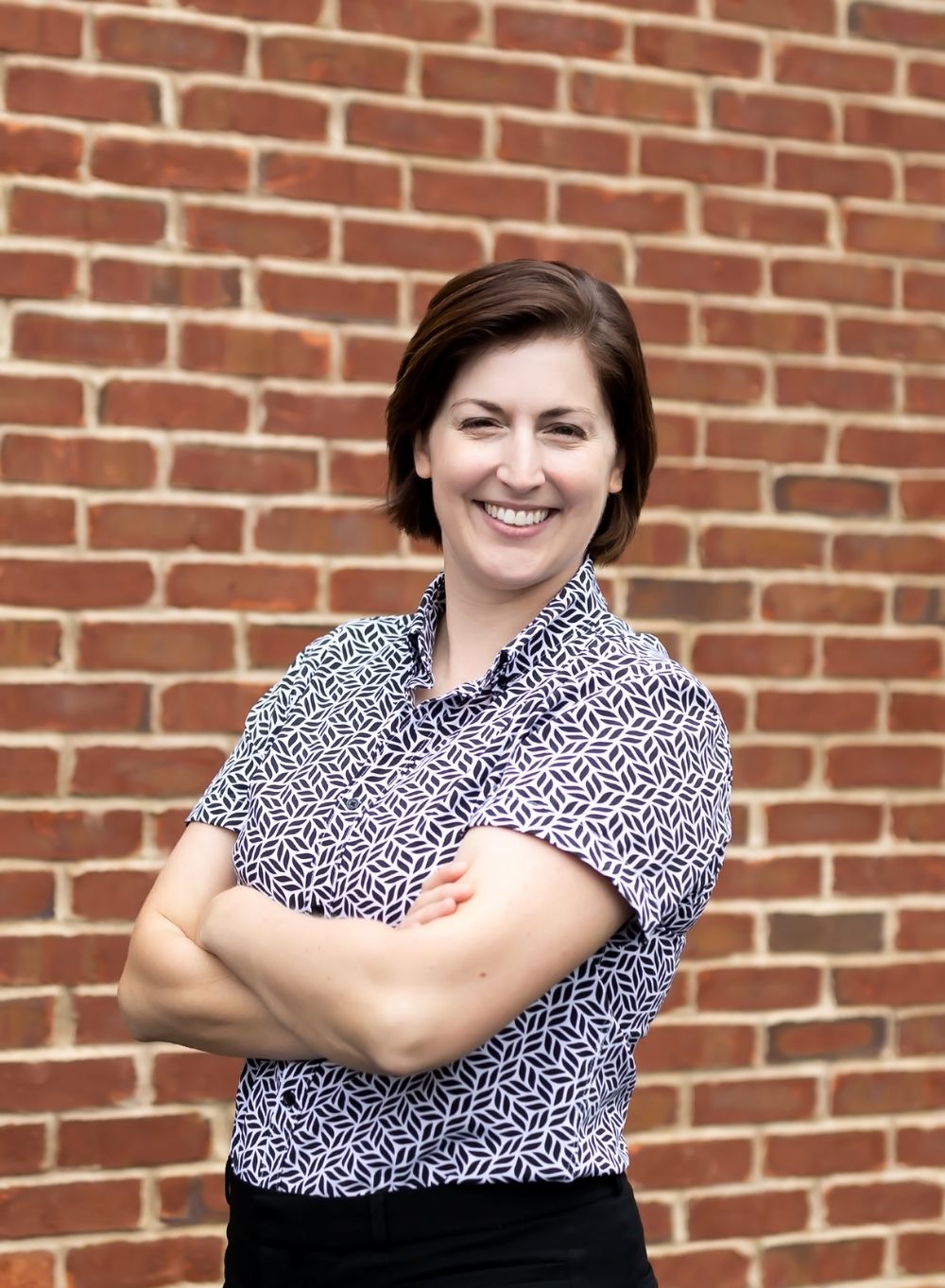 A person in a navy and white patterned top poses with crossed arms against a brick wall.