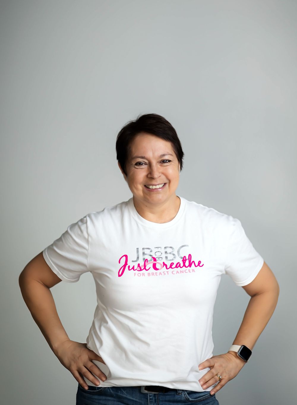 A person in a white t-shirt with pink text stands smiling against a gray background with hands on hips.