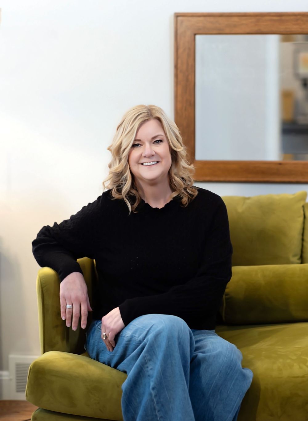 A person in a black sweater sits casually on a moss green velvet couch against a light colored wall with a framed mirror.