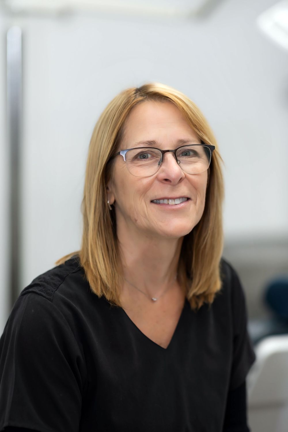 Professional wearing glasses and a black v-neck top smiles warmly in an office setting.
