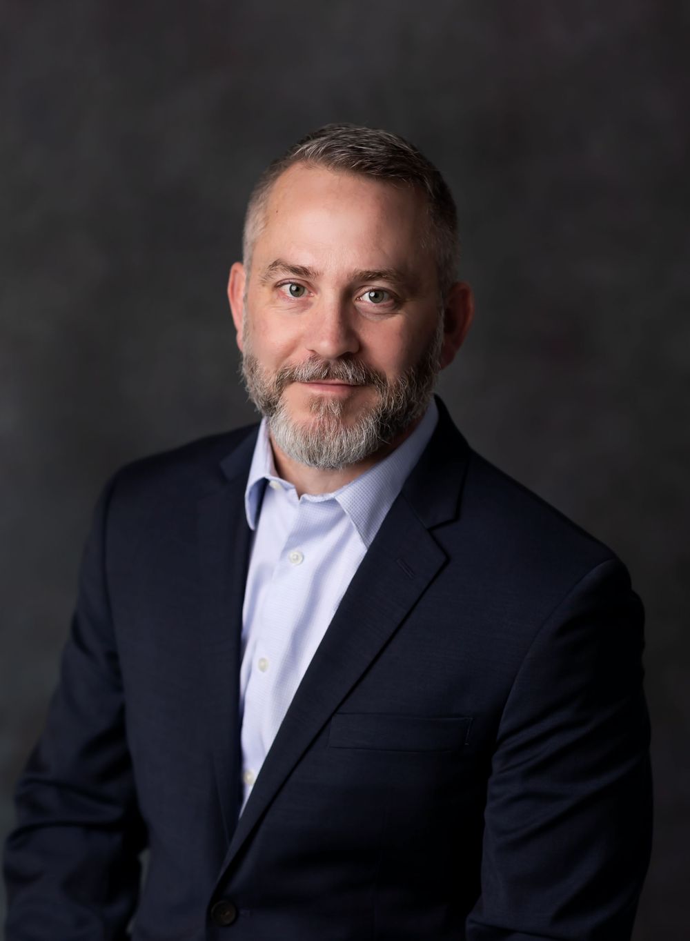 Professional headshot of a person in a navy suit jacket and light blue shirt against a dark gray background.