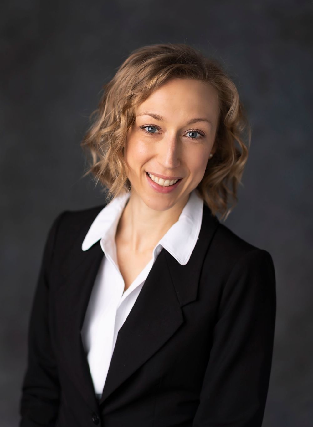Professional headshot of someone in a black blazer and white collared shirt against a dark background.