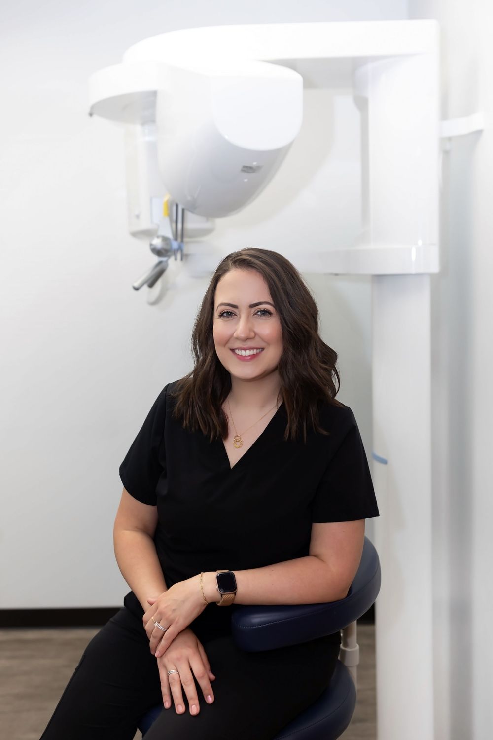 Professional wearing black scrubs sits in modern medical office with white walls and lighting fixtures.