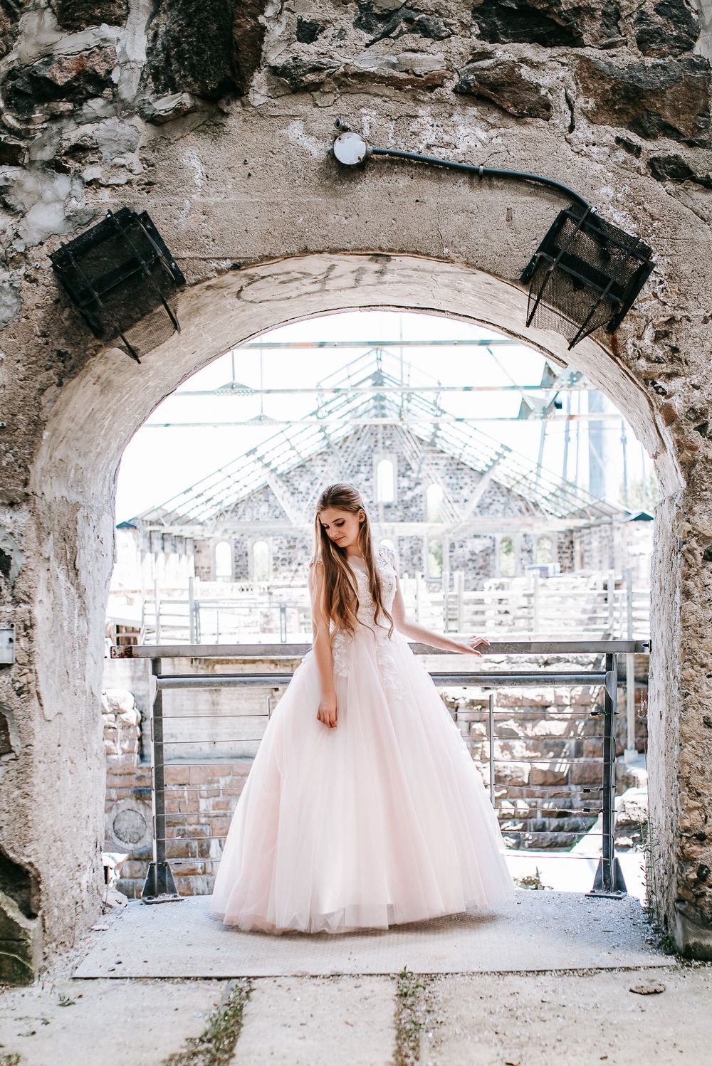 Ethereal ball gown showcased in rustic stone archway with greenhouse visible in background.
