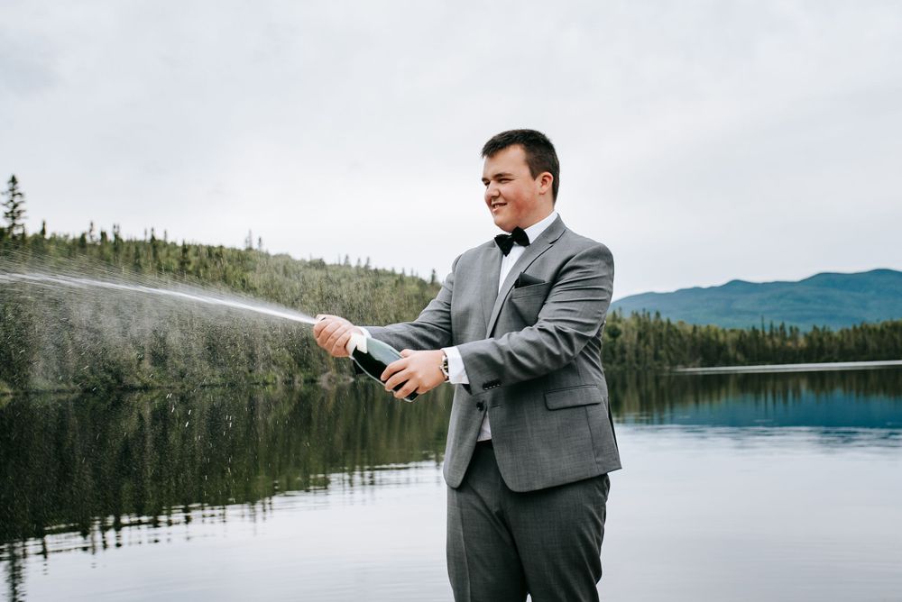 Person in grey suit stands against backdrop of calm lake and mountain scenery.