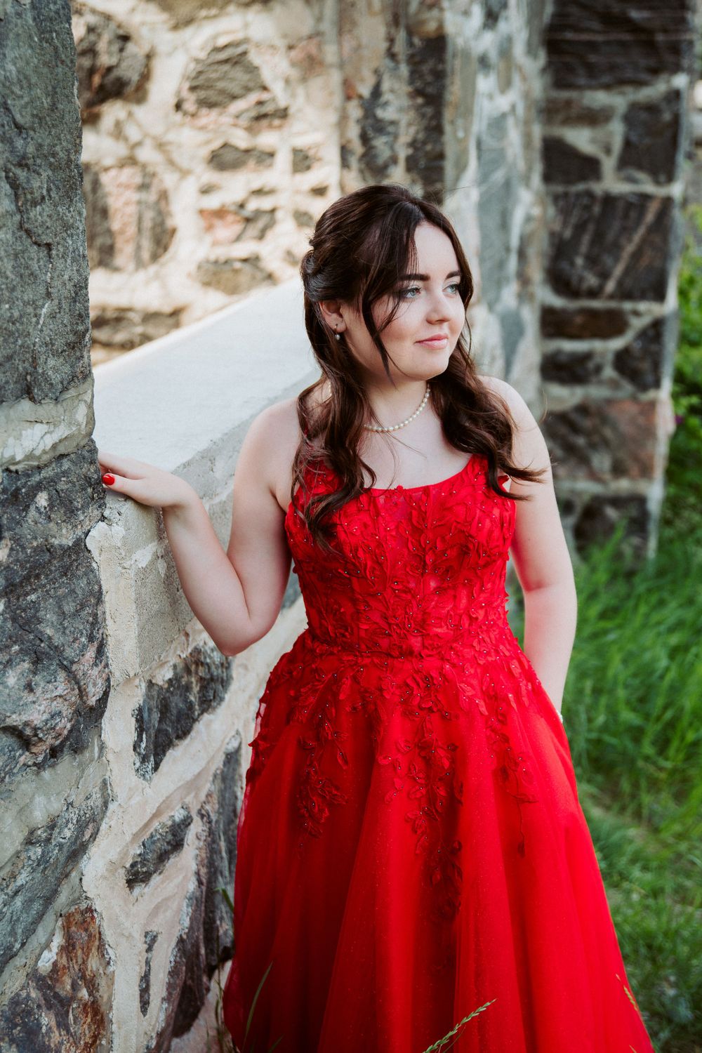 Woman in stunning red lace gown poses against stone wall in outdoor setting.