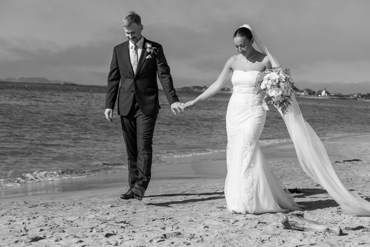 Bride and groom walking hand in hand along a sandy beach in black and white wedding photo.