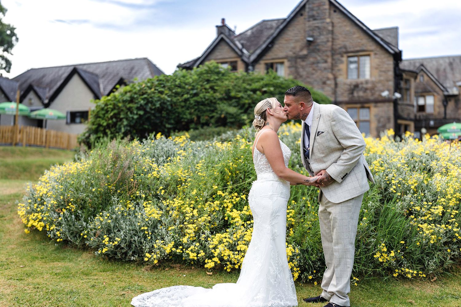 Bride and groom kissing in front of vibrant yellow flower garden outside a traditional stone building venue.