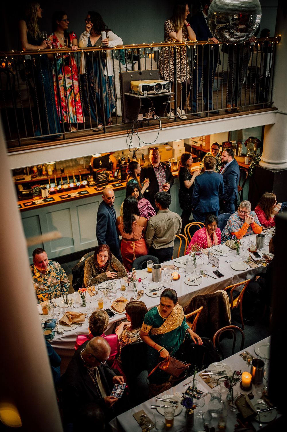 Overhead view of a lively restaurant scene with diners at long tables and spectators watching from a balcony above.