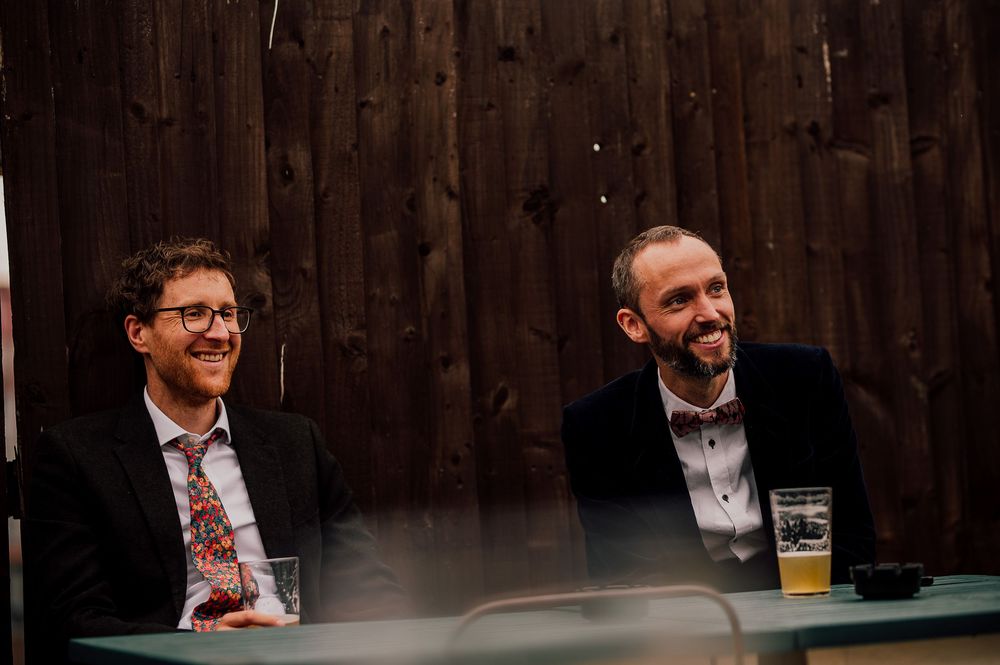 Two men in business attire share a laugh while sitting at a table with a beer glass in front of wooden wall.