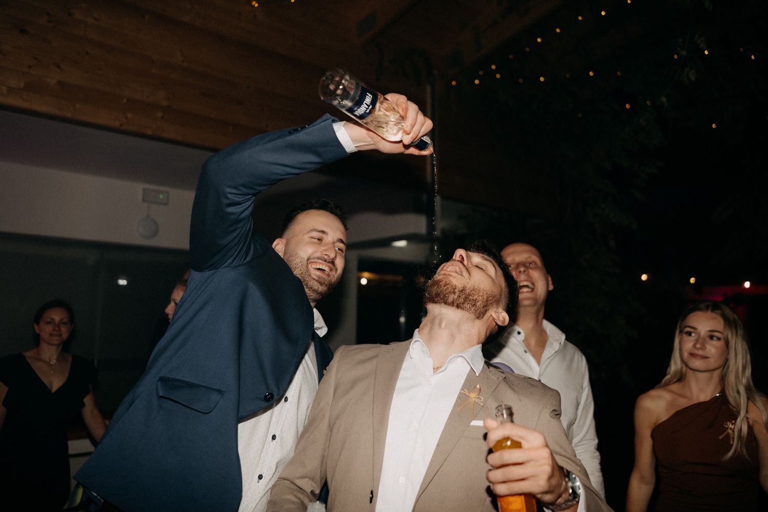 Two people in suits share a lively moment at a party while holding drinks.