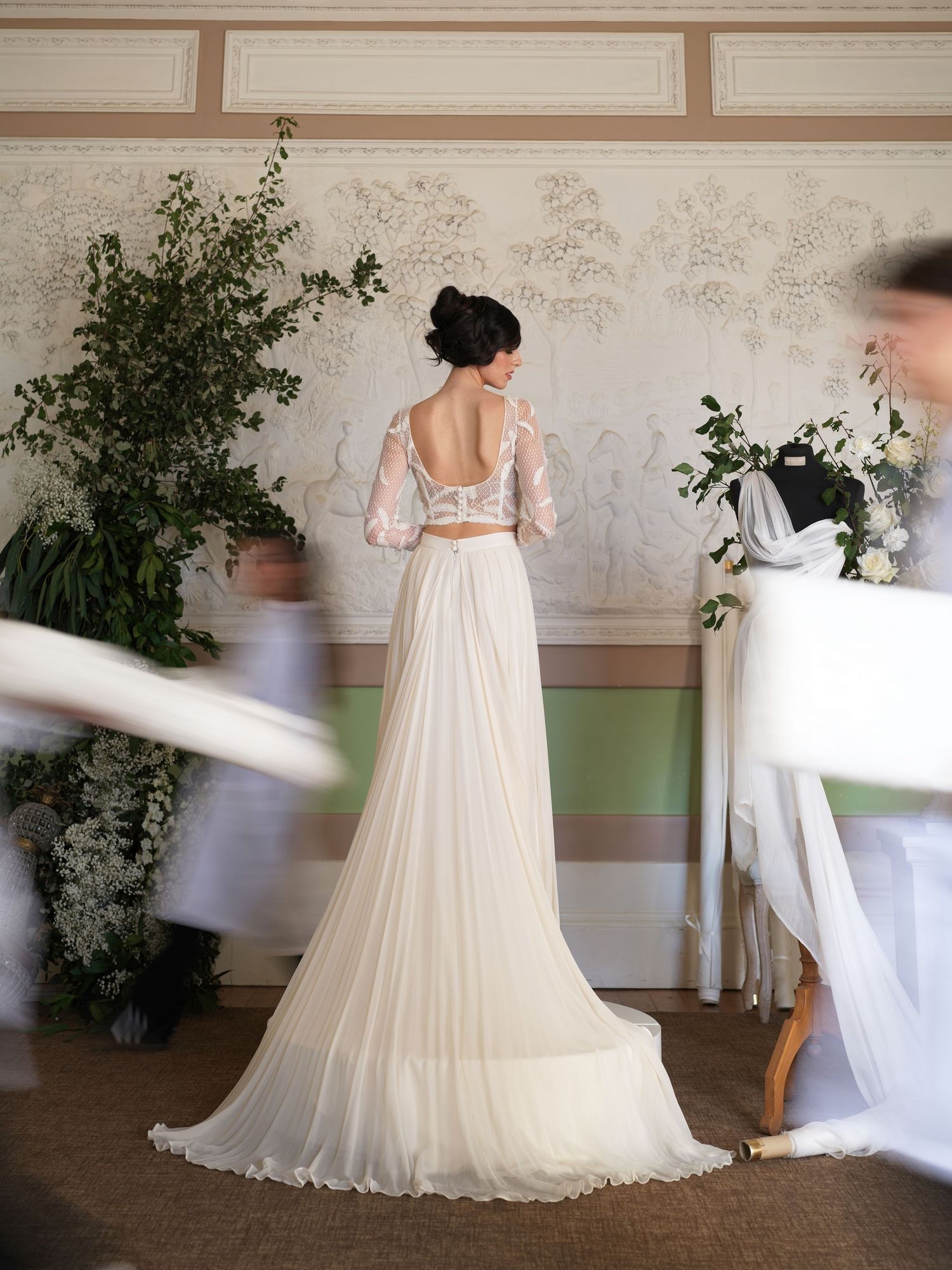 Elegant wedding dress with sheer lace sleeves and flowing tulle skirt shown from behind in a floral-decorated room.