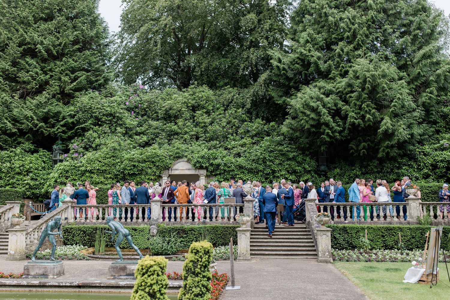 A group of people stand along a stone balustrade overlooking a garden in a park setting.