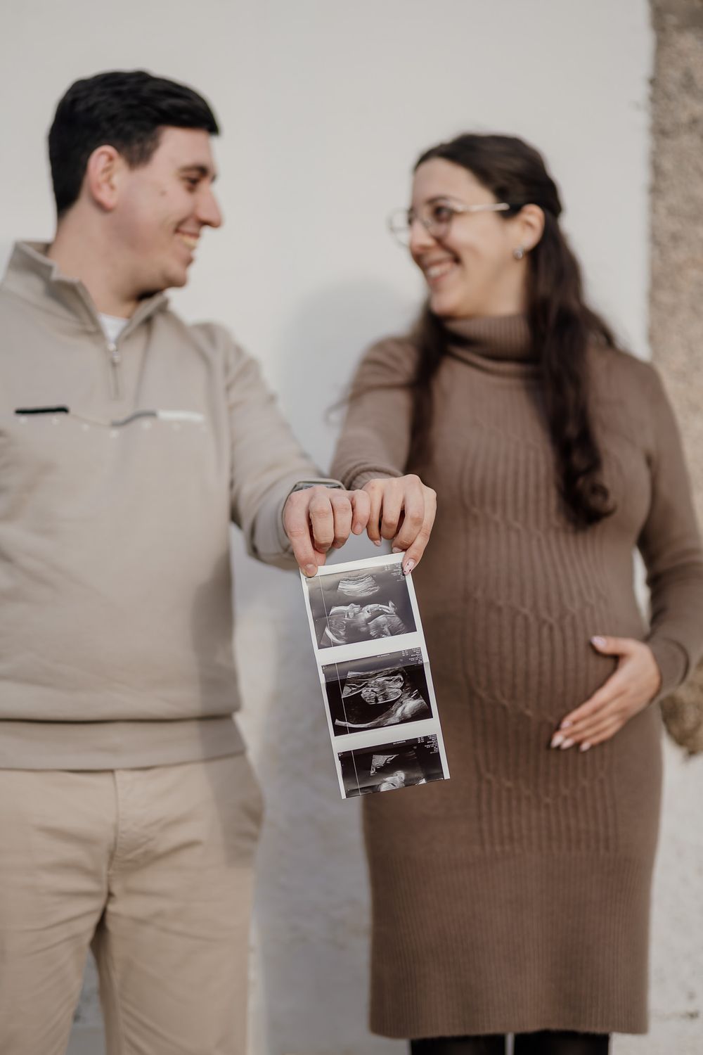 A couple in brown and beige clothing share a joyful moment while holding ultrasound photos in a pregnancy announcement series.