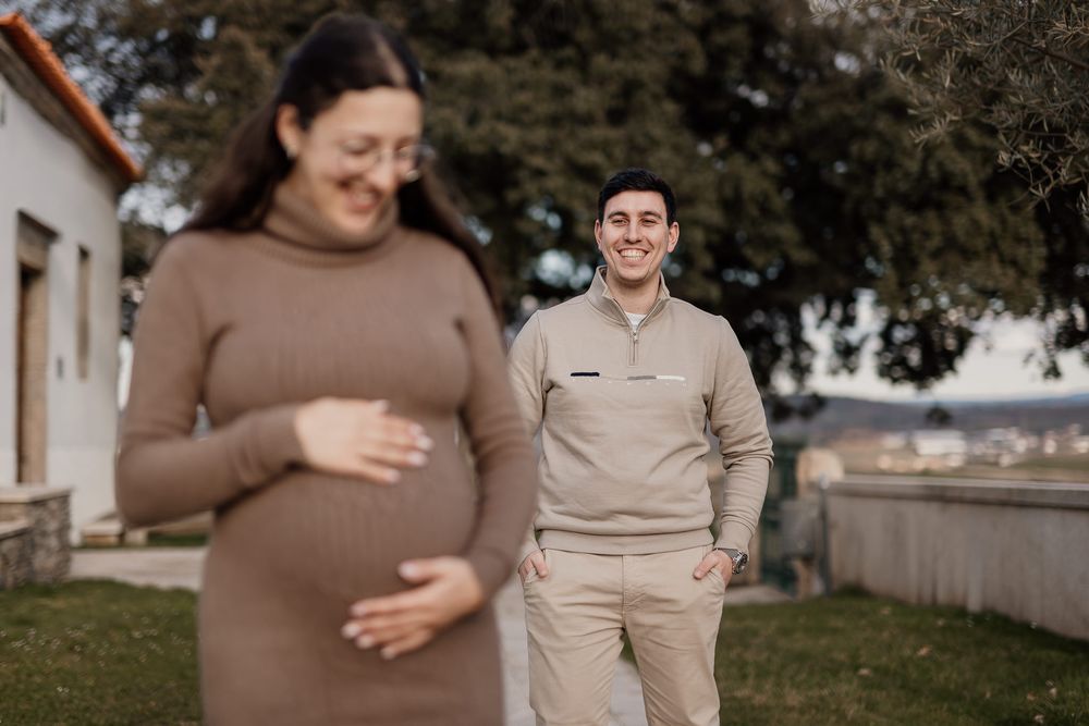 A couple in beige and brown outfits pose outdoors during a maternity photoshoot in a garden setting.