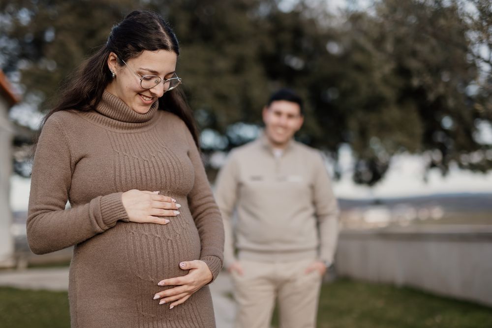 A pregnant woman in a brown sweater stands outdoors while touching her baby bump during a maternity photoshoot.