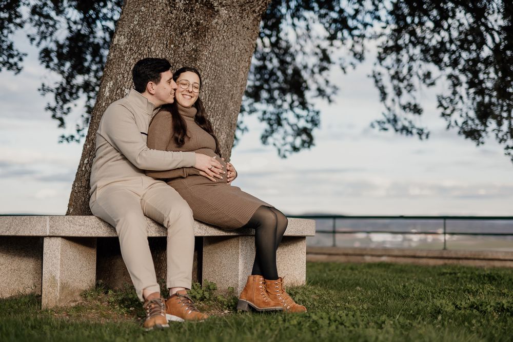 Couple in casual outfits sitting on wooden bench under tree branches during outdoor photo session.