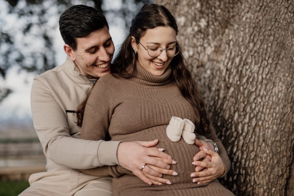 A couple in neutral toned sweaters embraces while standing near a tree trunk in an outdoor setting.