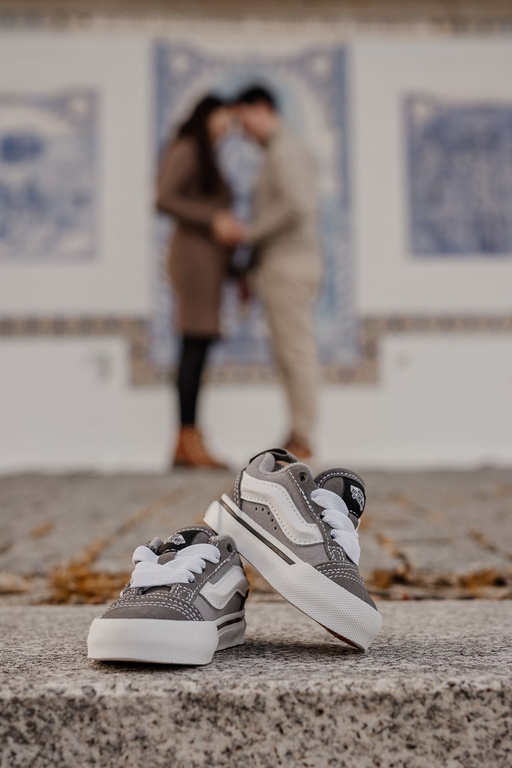 Small white and gray baby shoes placed on concrete ground with blurred figures in the background.