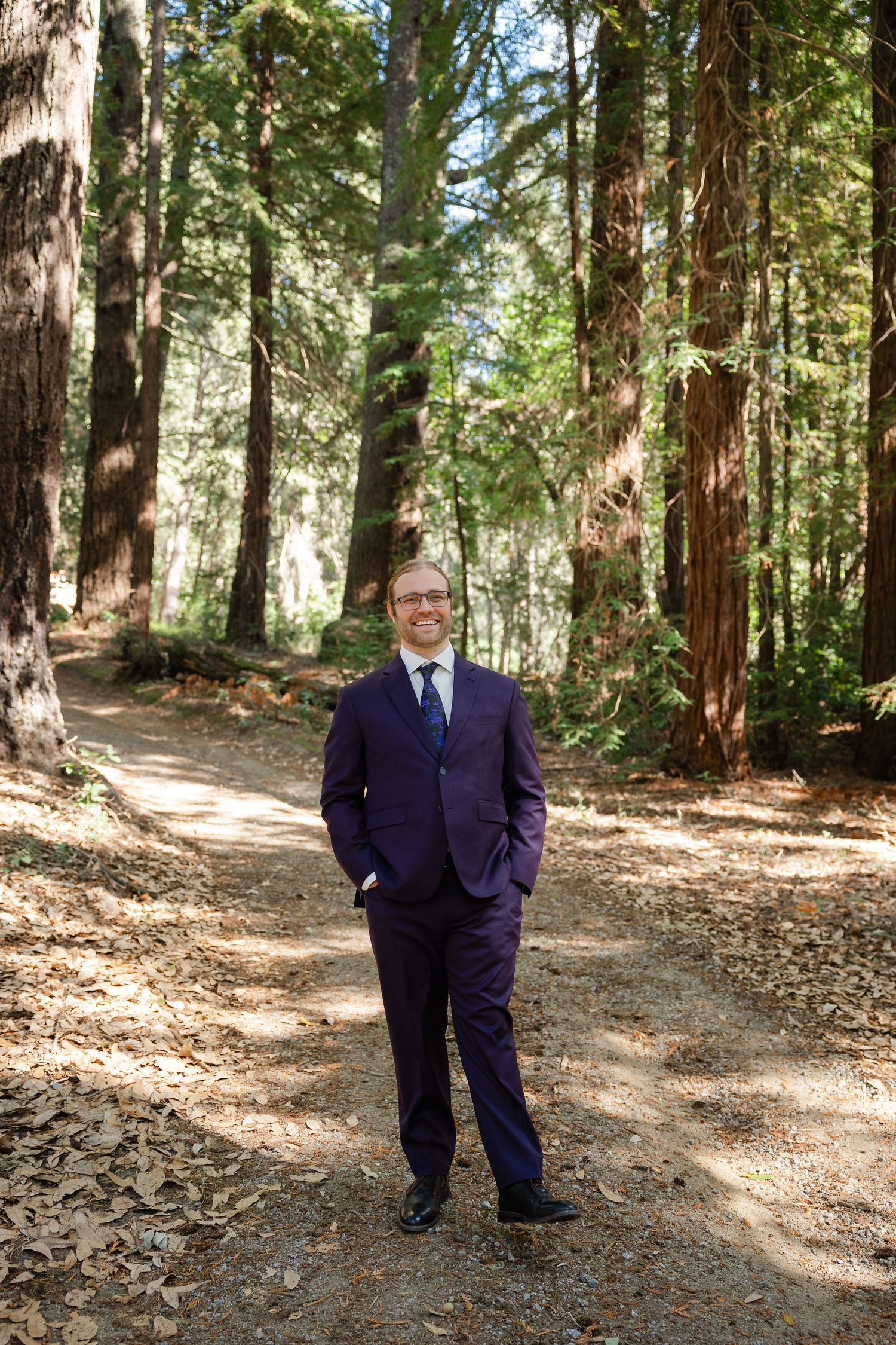 A person in a navy suit poses among towering redwood trees in a forest setting.