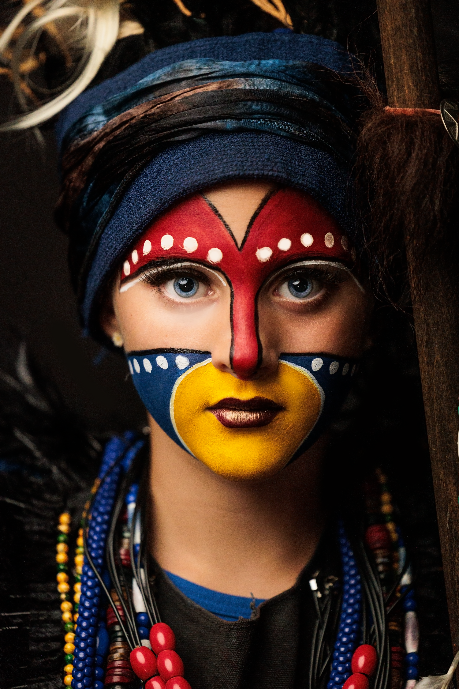 Woman with vibrant tribal face paint in red, blue, and yellow with beaded necklaces and headwrap.