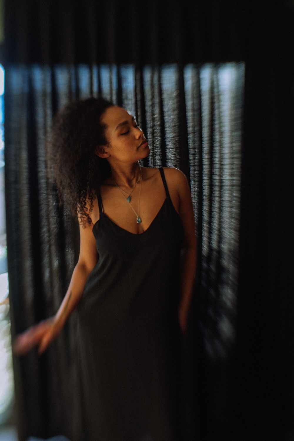 Moody indoor portrait with dark curtains and dramatic side lighting creating shadows.