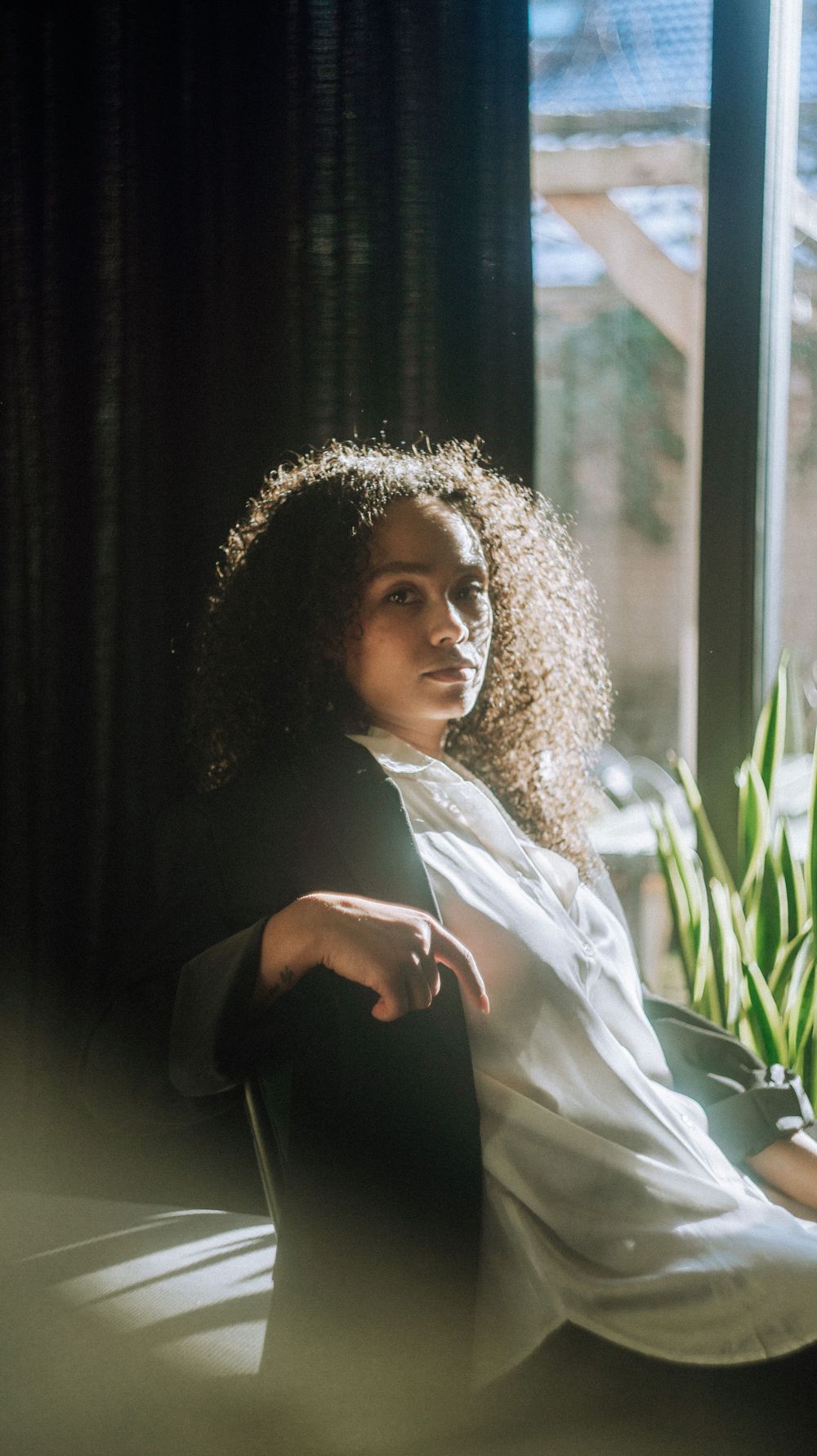 Relaxed portrait by a window with plants, wearing a white button-down shirt.