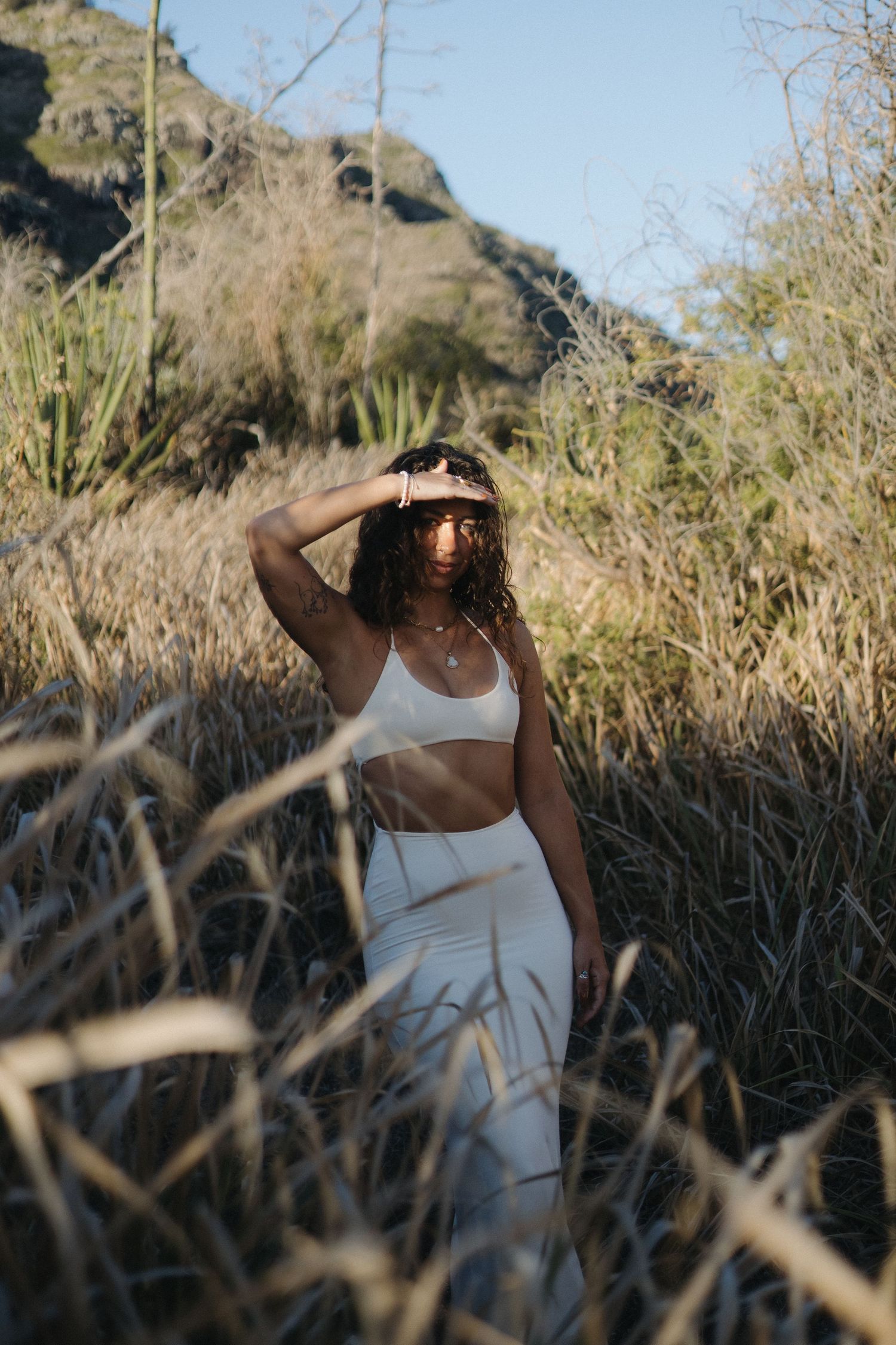 A person in white attire poses amid tall grass and desert brush on a sunny mountain trail.