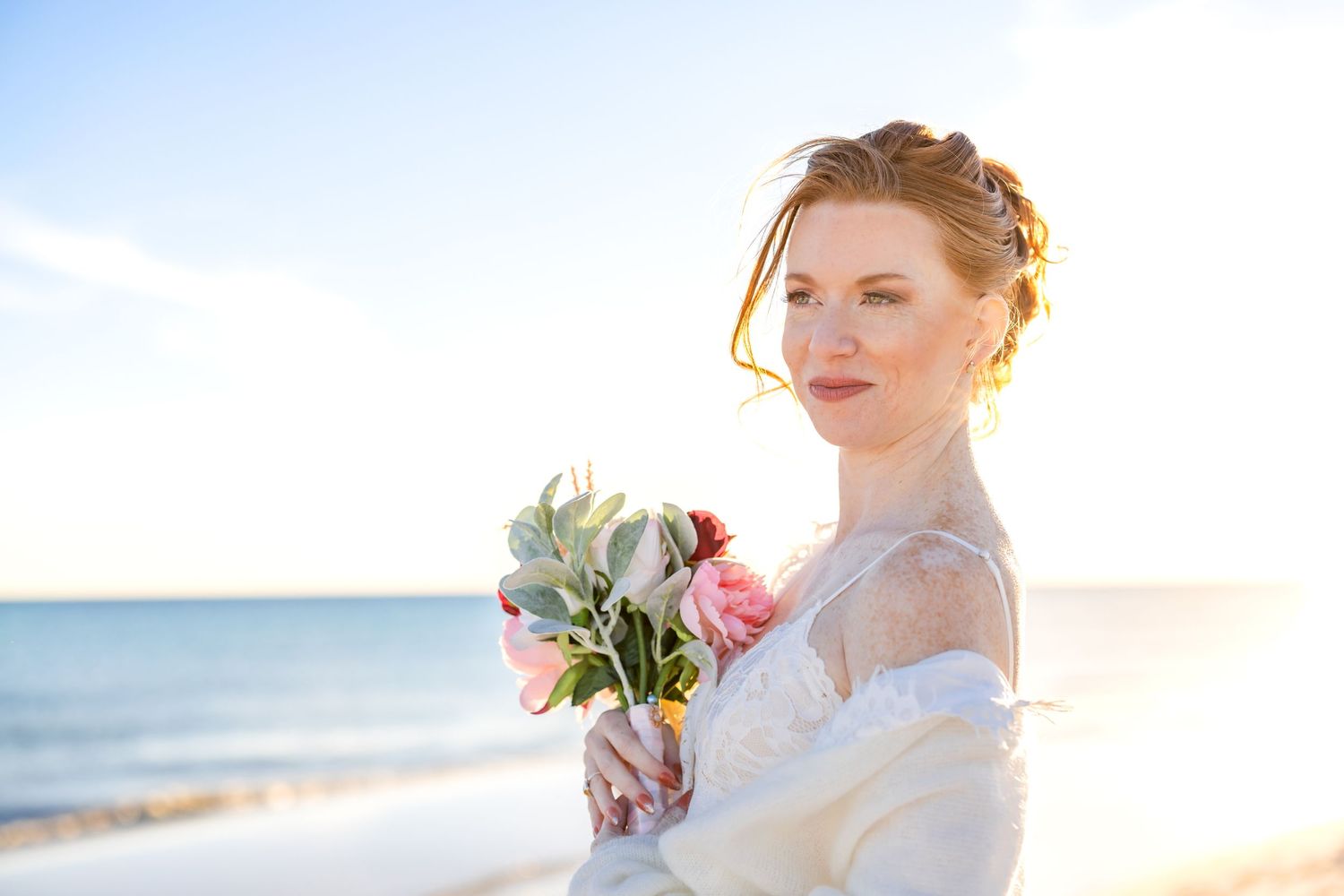 A wedding bouquet of pink and white flowers is held at a sunny beach during golden hour.